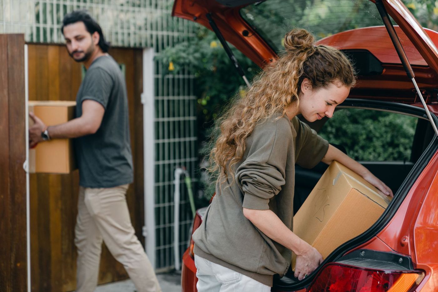 A woman lifting a cardboard box into the open boot of a car while a second person carries another box behind her