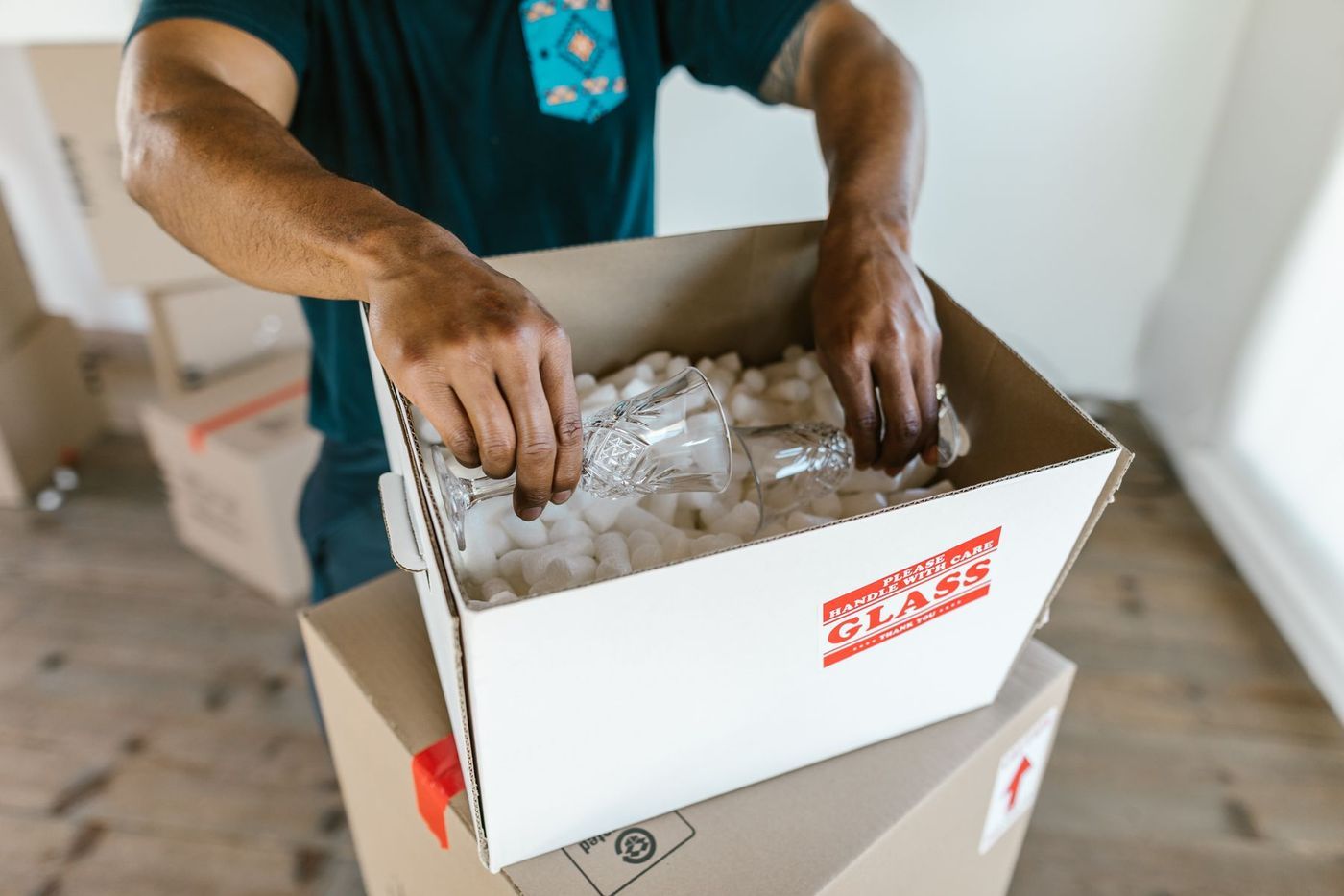 Hands lowering a wine glass into a box of packing chips, the box marked glass, handle with care