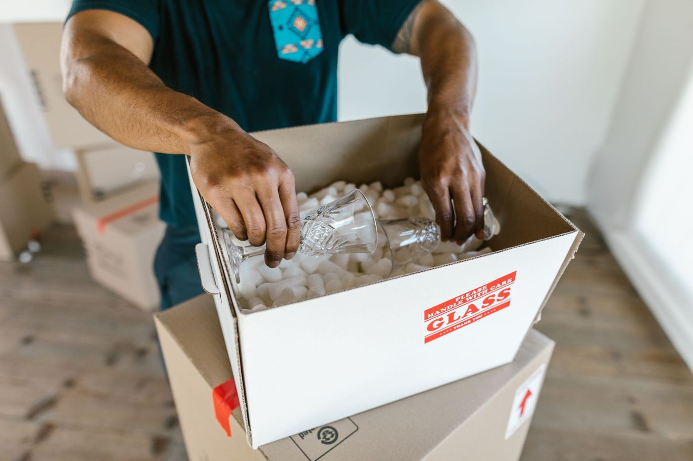 Hands lowering a wine glass into a box of packing chips, the box marked glass, handle with care